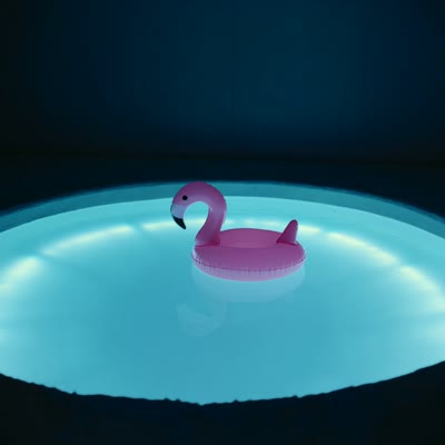 Slow handheld walks around the glowing pool, an inflatable flamingo drifts across, synth tone swells, a distant giggle. Audio: 90s camcorder hum, water lap, dreamy synth pad, distant crickets. Dreamcore liminal aesthetic.
