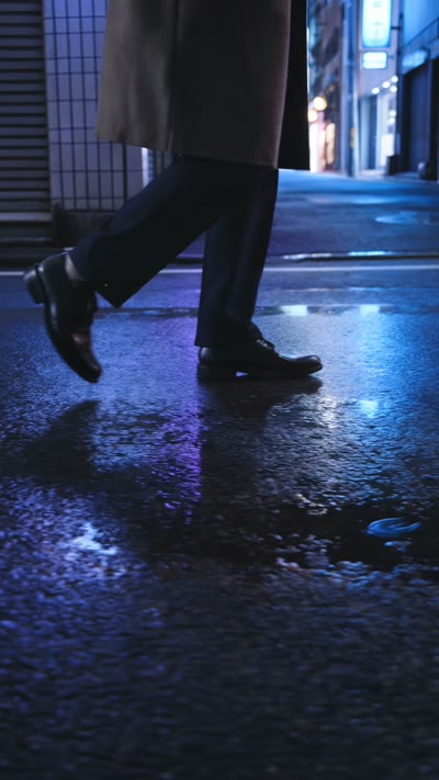 Side-tracking shot at ground level following the man as he walks through the rainy Tokyo alley at night, neon reflections streaking across wet asphalt, slow-motion rain, moody blue-and-purple lighting, cinematic film noir, 4K. Audio: rain, distant traffic, no dialogue.
